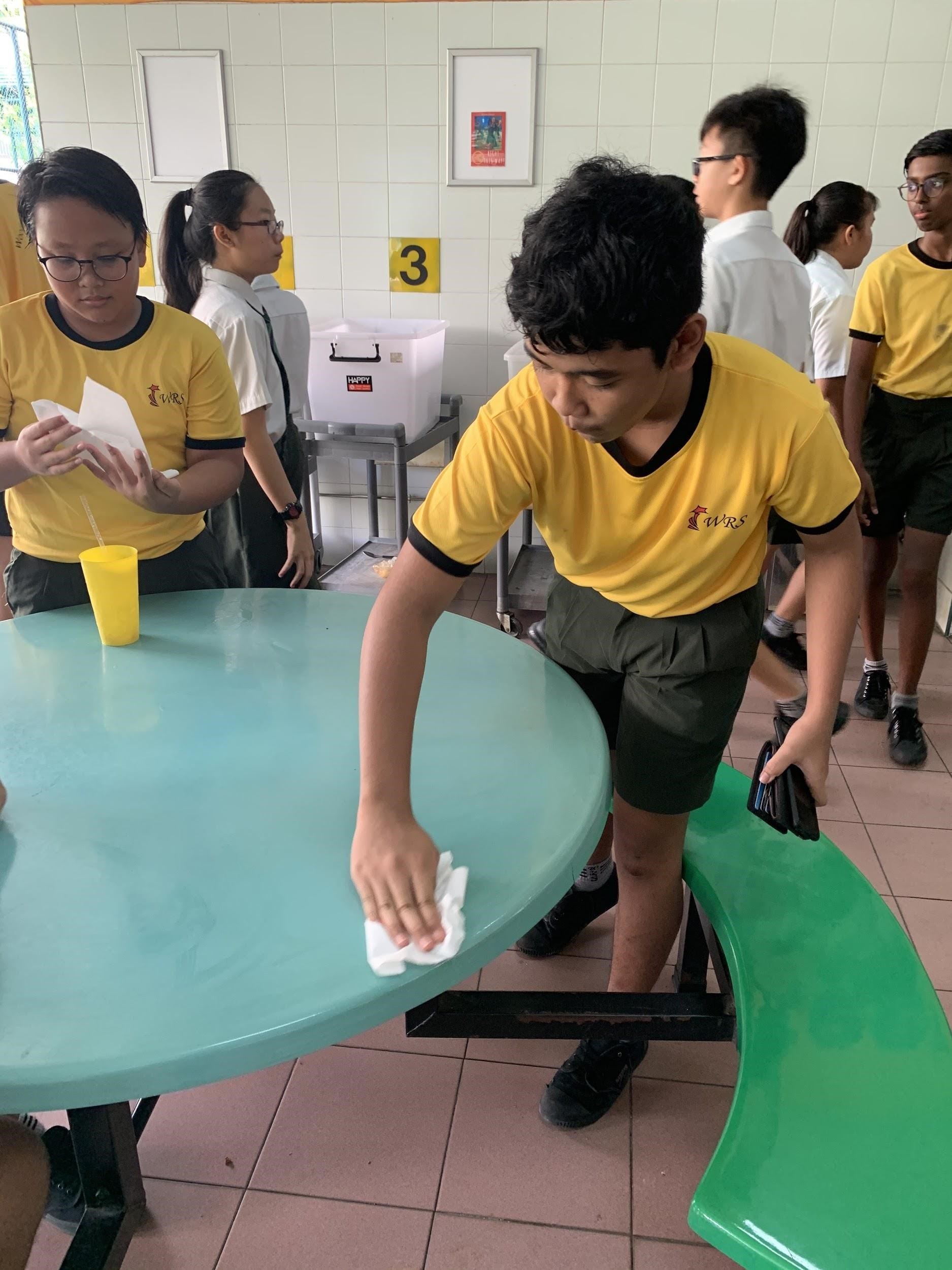 A student in a yellow WRS shirt wipes a round teal table with a cloth in a school canteen.